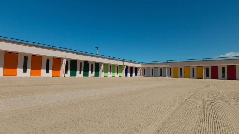 Rangée de cabines de plage colorées sous un ciel bleu, évocation des vacances et résidences secondaires.